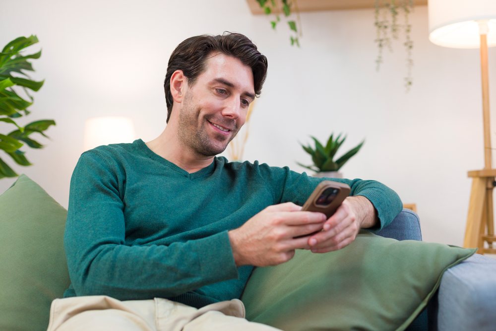 Man sitting on a couch at home using a smartphone in a relaxed indoor setting, technology, young adult.