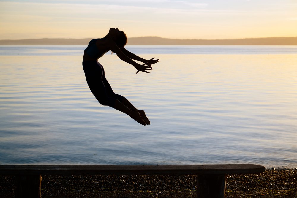 Woman jumping in an acrobatic pose over a bench near calm lake water at sunset, yoga, acrobatics, USA.