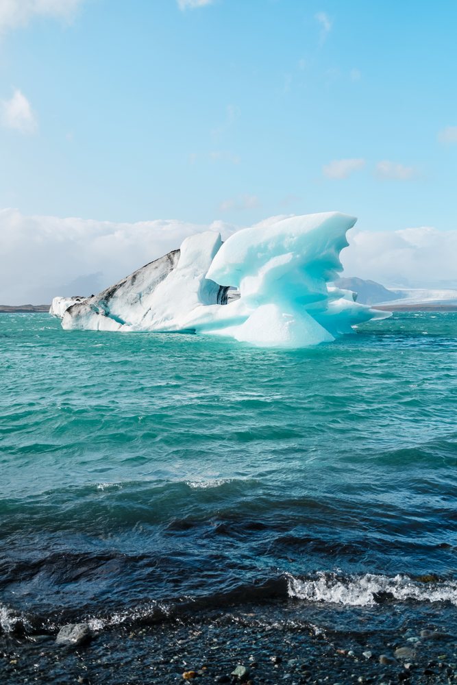 A large blue and white iceberg floats in icy water near a rocky shore under a partly cloudy sky, Iceland.