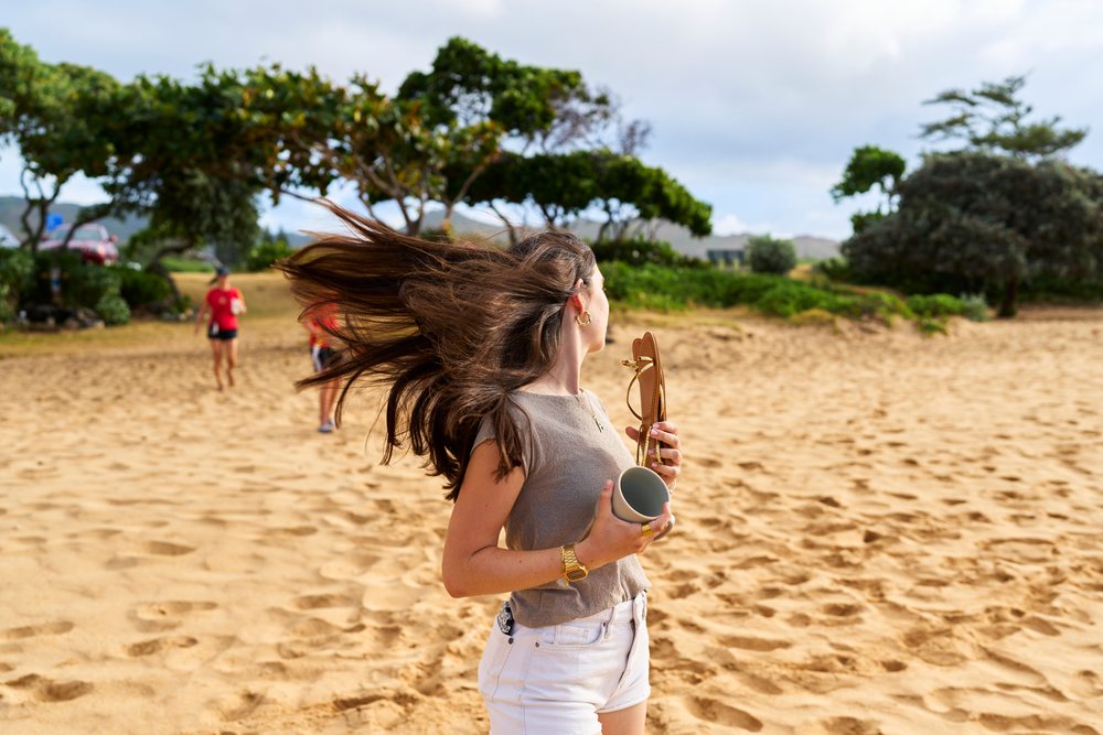 Woman flips her long brown hair while holding a sandal and cup on a sunny sandy beach with trees, Hawaii, USA.