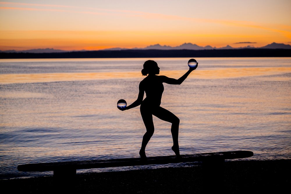 Silhouette of a woman practicing yoga and acrobatics balancing glass balls on a bench by a lake at sunset, USA.