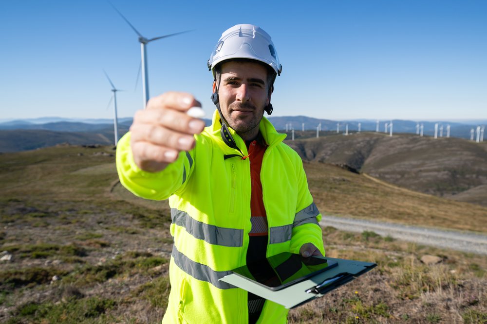 Man in helmet and high visibility jacket holding a tablet at a wind farm with turbines on hills, sustainability, renewable energy, Galicia, Spain.