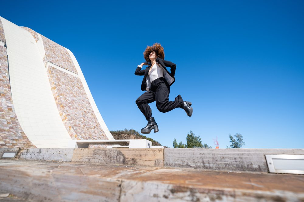 Businesswoman jumping energetically outdoors near a modern building under a clear blue sky in Santiago de Compostela, traveling executive, Spain, Antiago De Compostela.