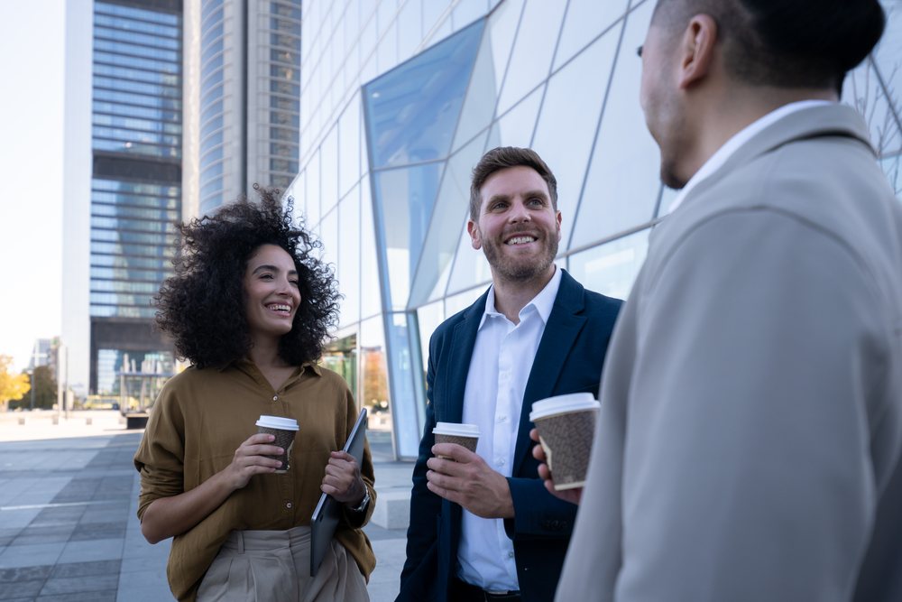 Three business colleagues converse and smile outdoors holding coffee cups near a modern office building, Madrid, Spain.