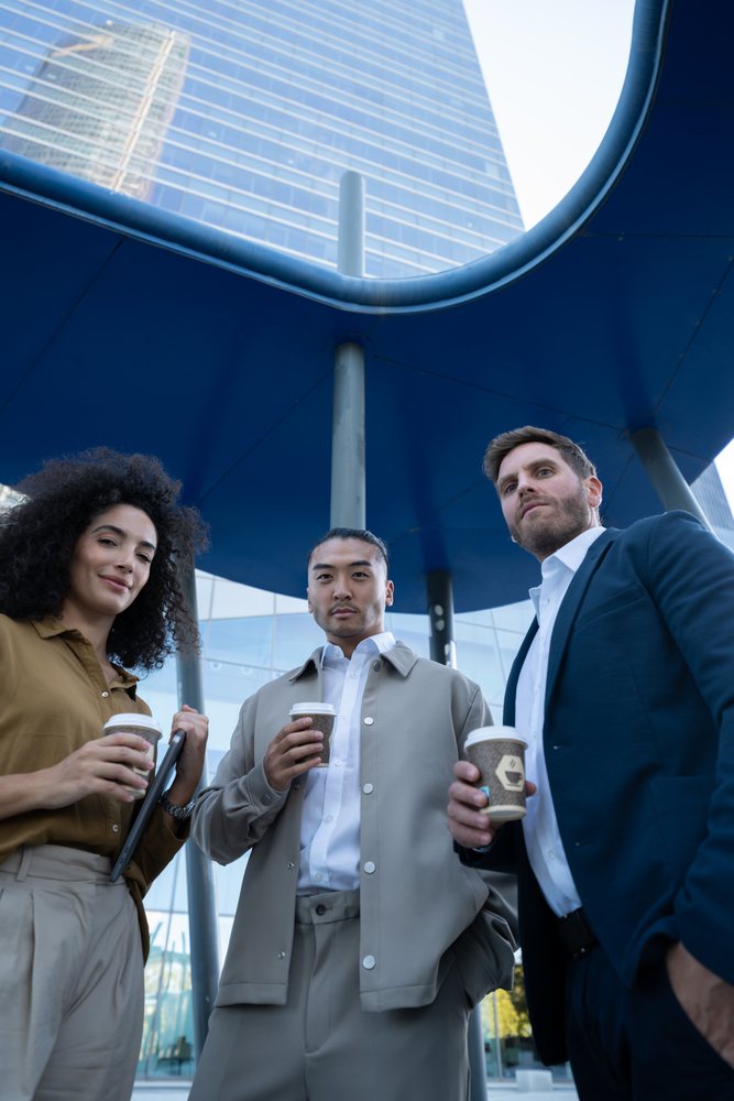Three businesspeople in formal and business casual attire hold coffee cups outdoors by a modern glass building in an urban setting, Madrid, Spain.