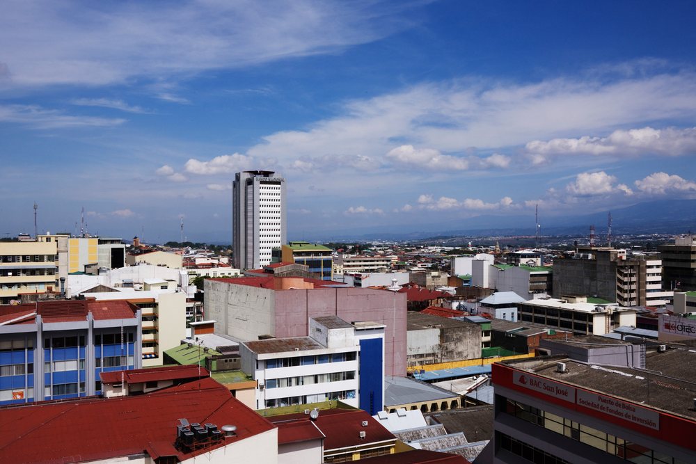 Cityscape of San Jose, Costa Rica showing urban buildings, a prominent tower, and rooftops under a blue sky with clouds during daytime.