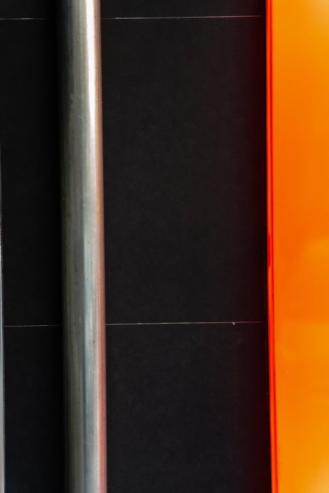 Vertical metal pipe mounted on a black tiled wall beside an orange panel creating industrial color contrast, texture, abstract texture, background, wallpaper, Spain.