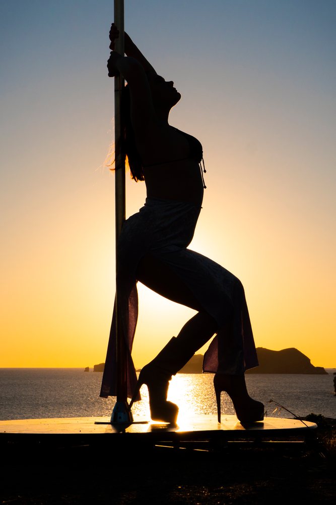Silhouette of a female pole dancer performing at sunset on a platform by the sea with islands in the background, pole dance, empowerment, Ibiza, Spain.