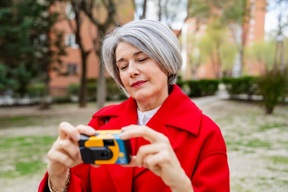 Woman with gray hair wearing a red coat takes a photo outdoors in an urban park with trees and grass around, Spain.