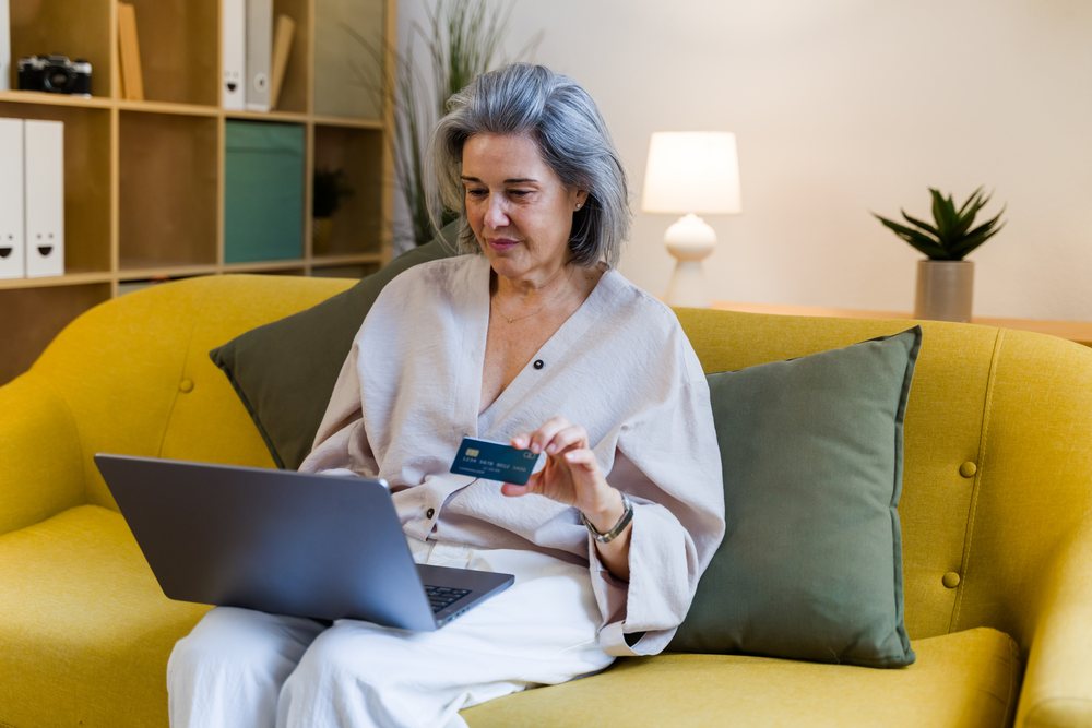 Woman with gray hair sits on a yellow sofa indoors using a laptop and holding a credit card, Spain.
