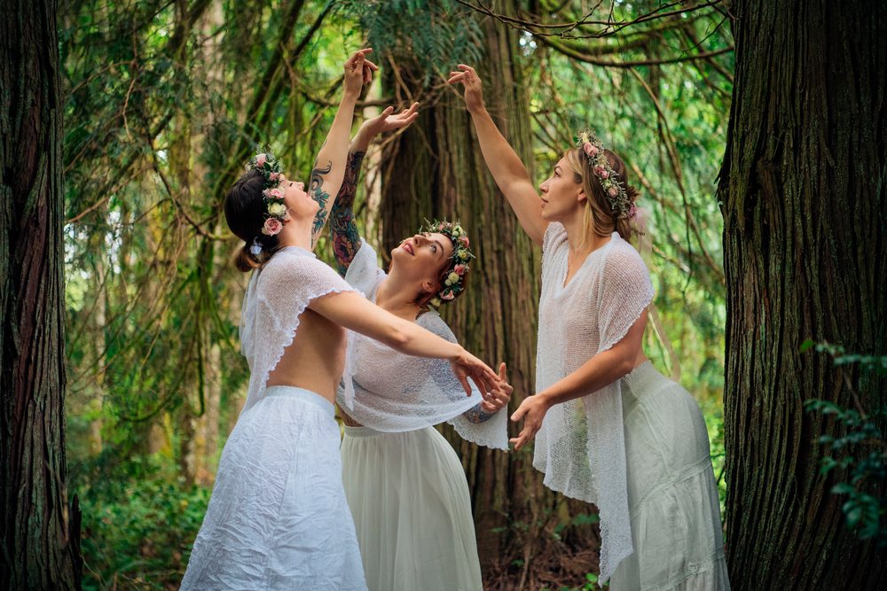 Three women wearing white shawls and flower crowns dance gracefully in a wooded forest setting, muses, USA.