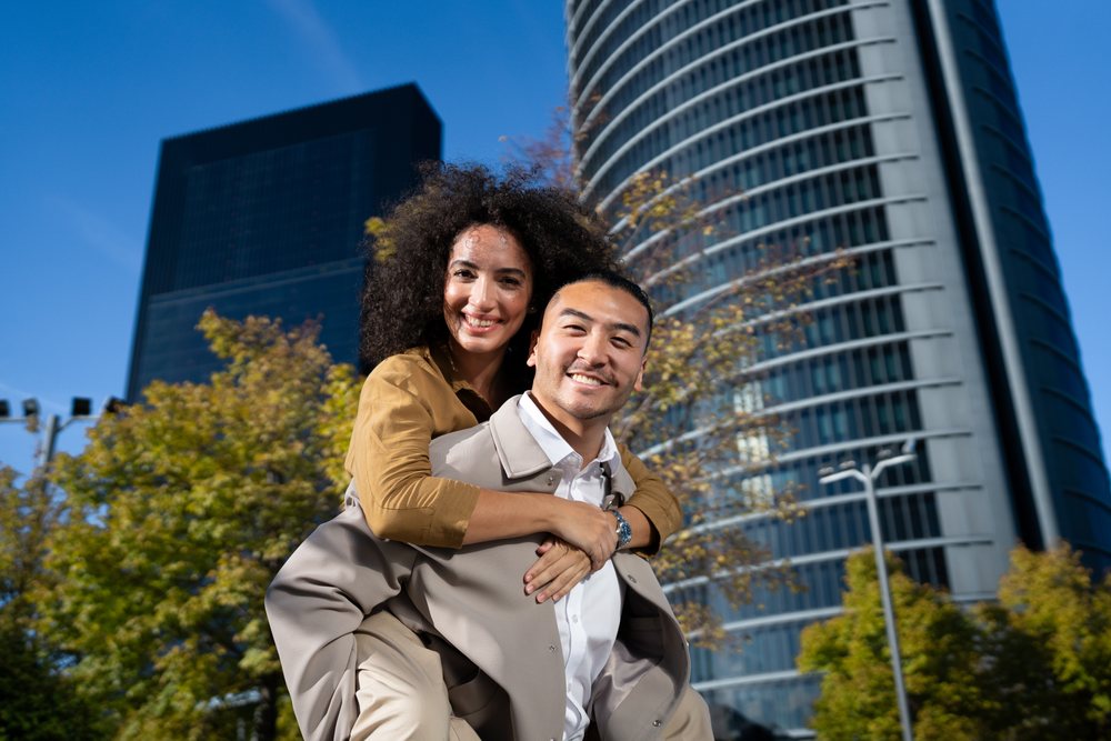 Man giving piggyback ride to woman outdoors in an urban area with modern buildings and trees under blue sky, Madrid, Spain.