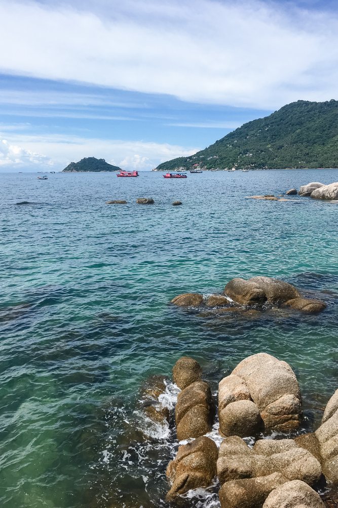 Rocky shore with waves, boats, and distant islands under a blue sky, Philippines.