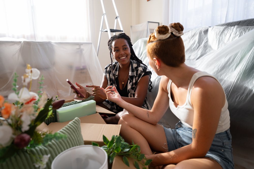 Two women happily pack boxes and chat during moving day in a bright home interior covered with plastic sheets, Spain.