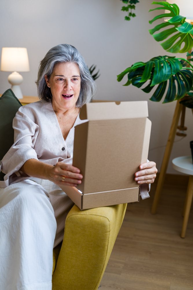Adult woman with gray hair sitting in a yellow armchair opening a brown cardboard box in a living room with a white lamp and indoor green plant with large leaves, Spain.