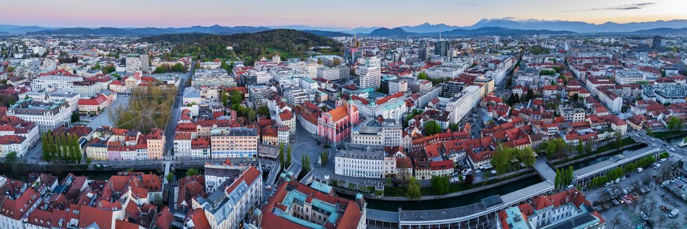 Aerial view of Ljubljana featuring the river, multiple bridges, red-roofed buildings, and mountain range at sunset, landscape, cityscape, Slovenia.