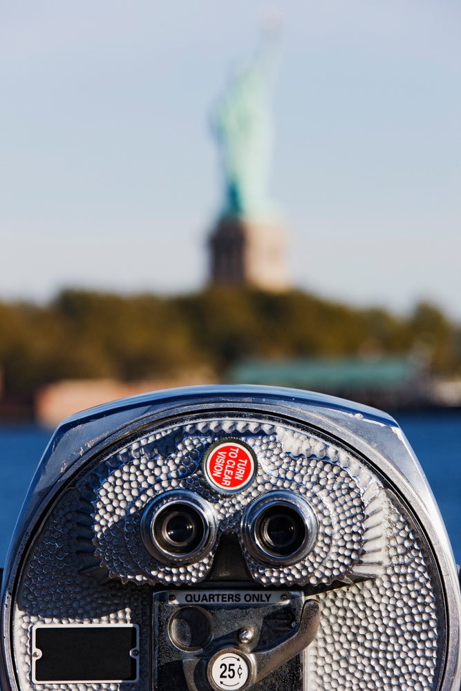 Coin-operated binoculars with a red label and Statue of Liberty blurred across the water on Liberty Island, New York, USA.