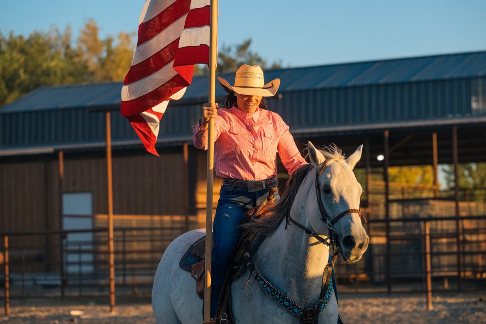 Woman wearing a cowboy hat rides a white horse holding an American flag outdoors at a Southwest USA ranch during sunset.