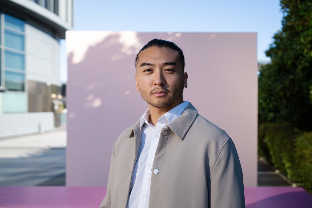 Man wearing beige jacket and white shirt stands outdoors in an urban setting with greenery and clear sky, Madrid, Spain.