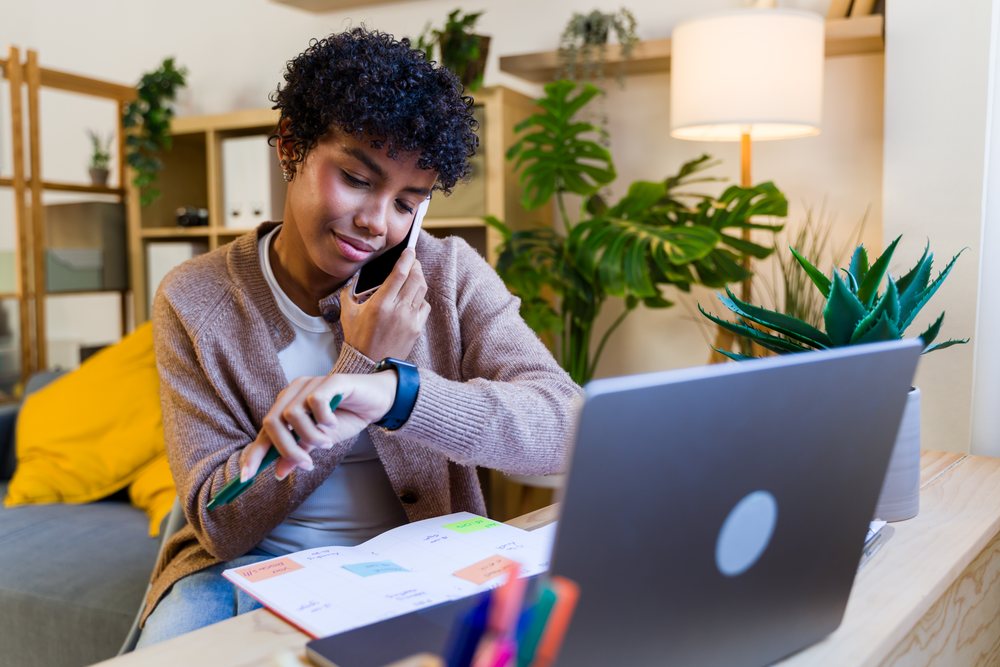 Woman checking her watch while talking on the phone at a home office desk with a laptop, planner, and indoor plants nearby, Spain.