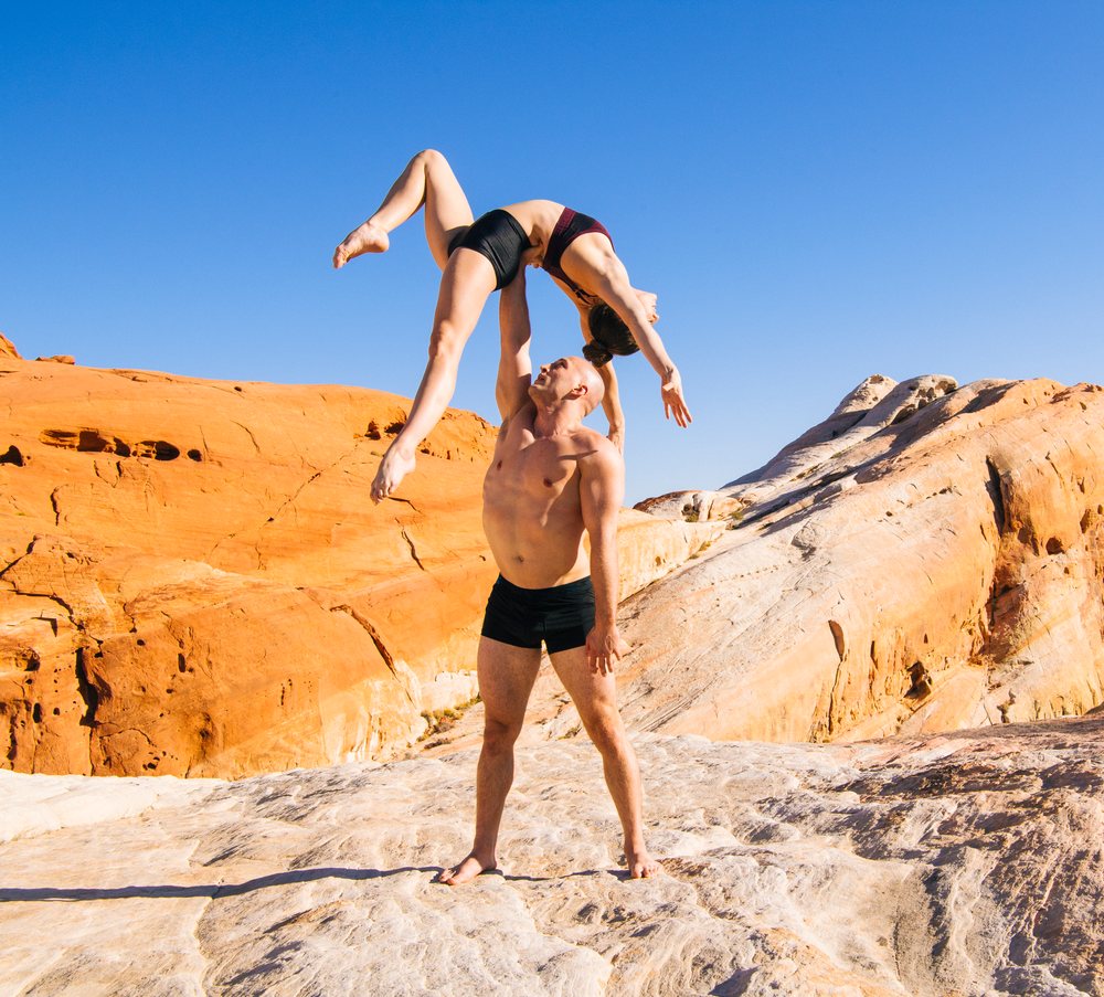Two barefoot athletes perform an acrobatic yoga pose on rock formations in a USA desert under a clear blue sky.