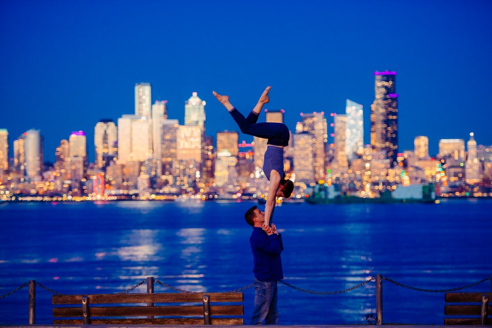 Two people perform a handstand partner acrobatics yoga pose on benches by the Seattle waterfront at night with the illuminated city skyline in the background, USA.