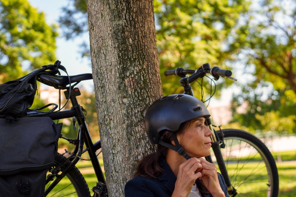 Woman puts on a black helmet while leaning against a tree with a bicycle behind her in a park in Málaga during daylight.