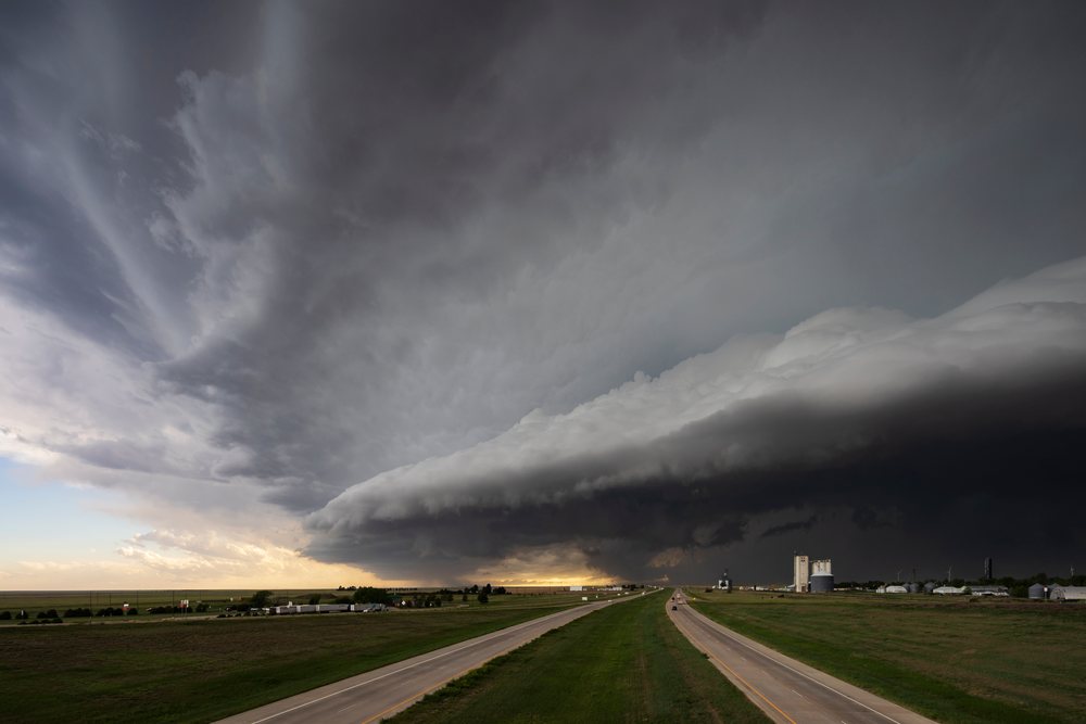Supercell storm clouds dominate the sky over a two-lane highway crossing open grassy fields and rural silos in Tornado Alley, Storm Chasing, Weather, Environmental Issues, Arriba, Colorado, USA.
