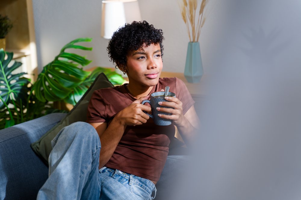 Woman with curly hair holds a grey mug with a spoon while sitting on a sofa in a modern, cozy living room with indoor plants and soft natural light, Spain.