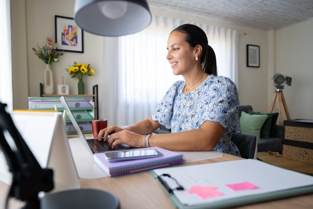 Woman typing on a laptop at a desk in a home office with organized stationery and flowers, telecommuting, Spain.