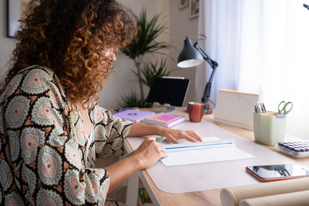 Woman with curly hair measuring white paper with a ruler on a desk in a cozy home office with natural light and organizing tools, Spain.
