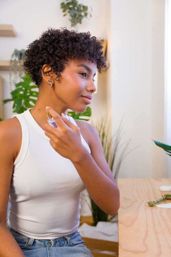 Young woman with curly hair sitting at home applying perfume near neck in a bright room with indoor plants and wooden table.