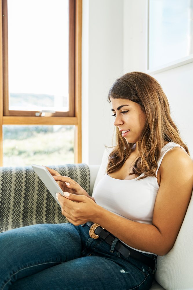 A white woman sits on a couch using a tablet in a bright home interior