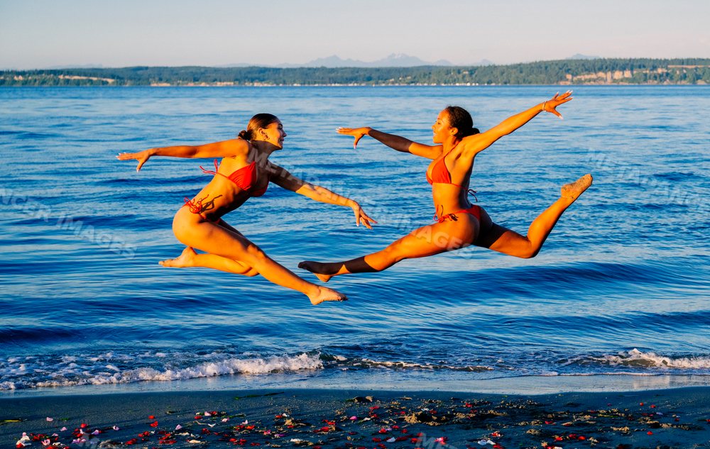 Two women jump in unison on a beach shoreline in WA, USA
