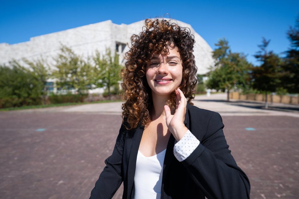 Businesswoman with curly hair wearing a black blazer and white blouse smiling outdoors with a modern building and trees in the background, S, traveling executive, Spain, Antiago De Compostela.