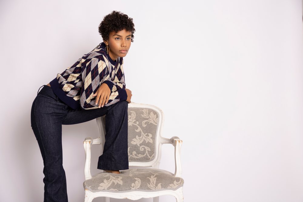 Young woman wearing an argyle sweater and dark jeans leans thoughtfully on a vintage upholstered armchair in a studio setting, fashion, young culture.