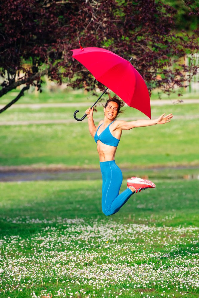 Joyful woman in blue sportswear jumps outdoors holding a red umbrella on a sunny grassy field, Calisthenics, USA.