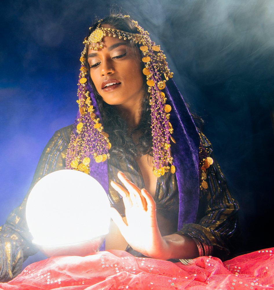 A woman dressed as a fortune teller with a purple veil and gold coin jewelry gazes into a glowing crystal ball in a smoky, mystical indoor setting in the, USA.