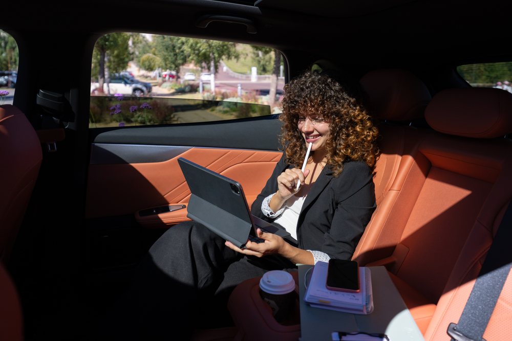 Woman working with a digital tablet and pen inside a car backseat with a notebook and coffee cup beside her in daylight, remote work, businesswoman, Spain.