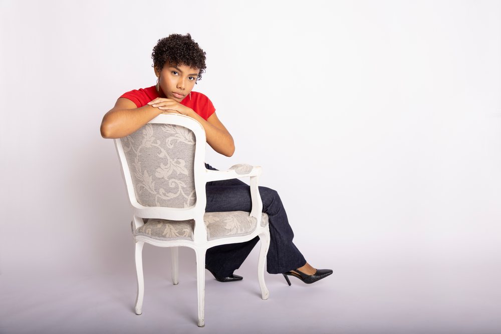 Young woman with curly hair wearing a red t-shirt, dark jeans, and black heels sits backwards on a white vintage chair in a minimalist studio setting, fashion, clothes, young culture.