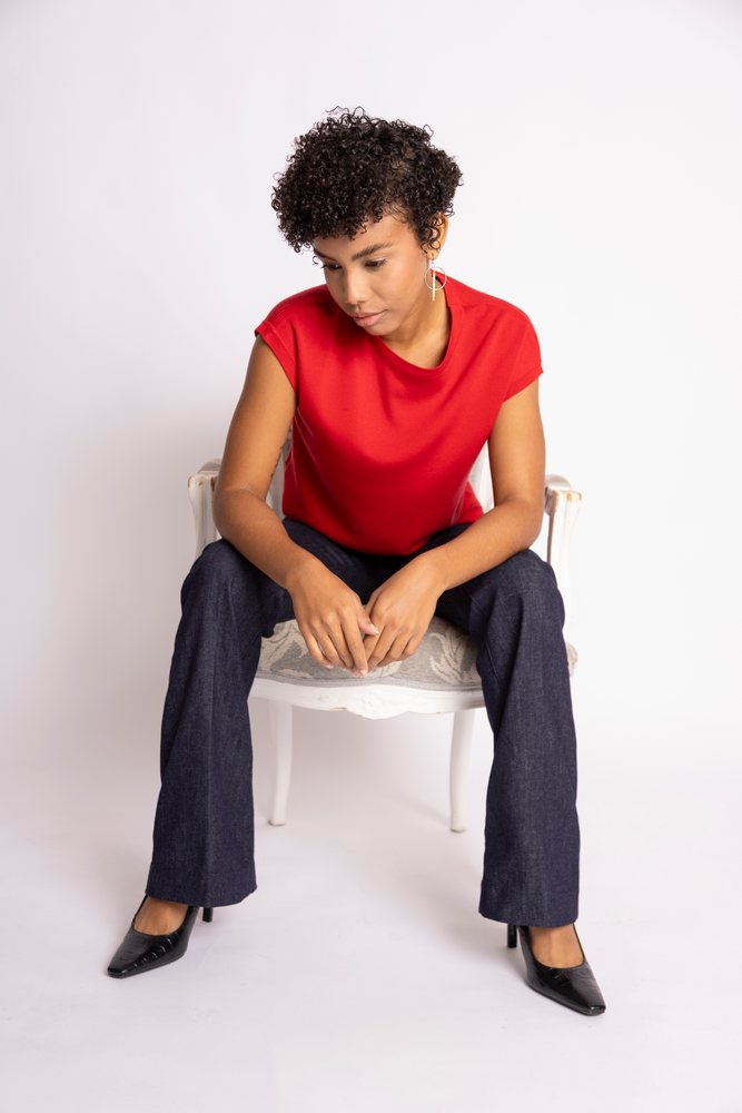 Woman with curly hair wearing a red shirt and dark jeans sits on a white chair in a studio setting, fashion, clothes, young culture.