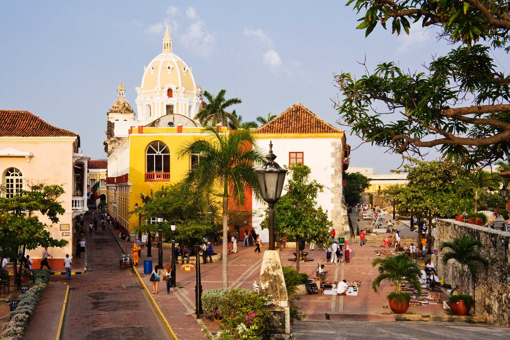People walking and gathering in a plaza with colorful colonial buildings and historical architecture, Cartagena, Colombia.