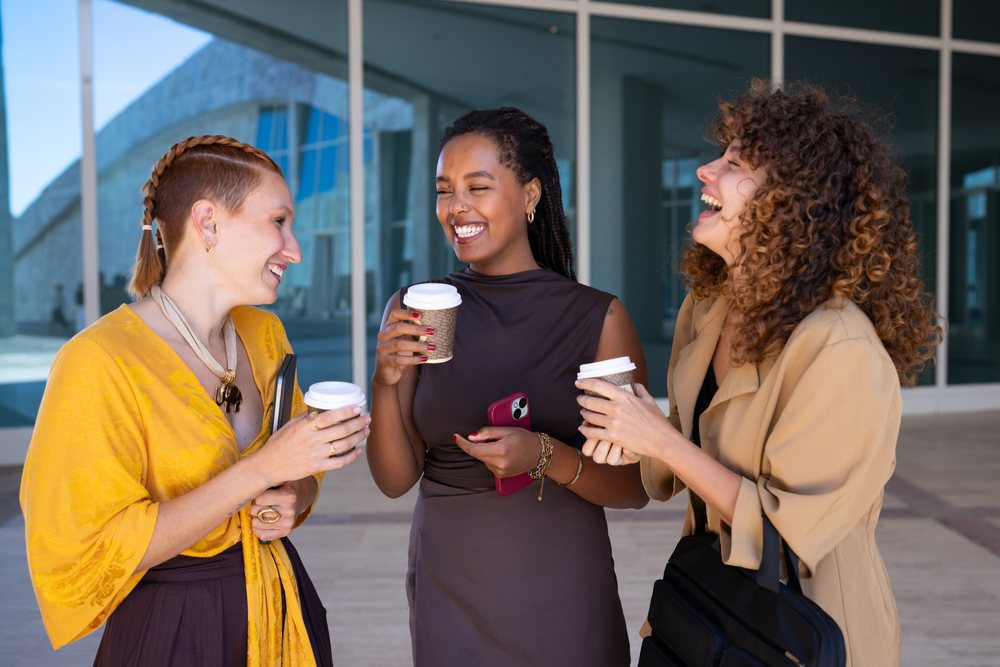 Three women laughing and holding disposable coffee cups outside a modern glass building in Spain during a casual conversation.