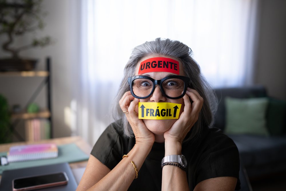 Older woman with gray hair and black glasses seated indoors at a table wearing URGENTE and FRÁGIL stickers on her forehead and mouth, Spain.