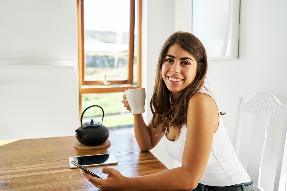 A young white woman smiling and holding a mug while sitting indoors