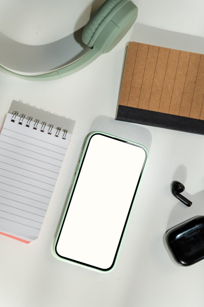 Smartphone with blank screen on white desk alongside headphones, wireless earbud, notepad, and black earbud case,, white screen, Spain.