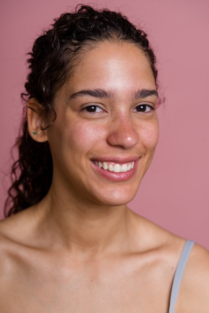 Smiling woman with curly hair and freckles posed against a pink studio background in a beauty shoot, Spain.