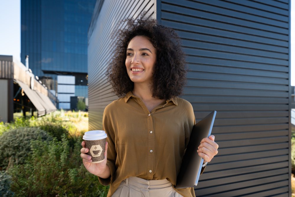 Smiling woman with curly hair holding a disposable coffee cup and closed laptop stands outdoors by a modern building, on a clear daylight, Madrid, Spain.