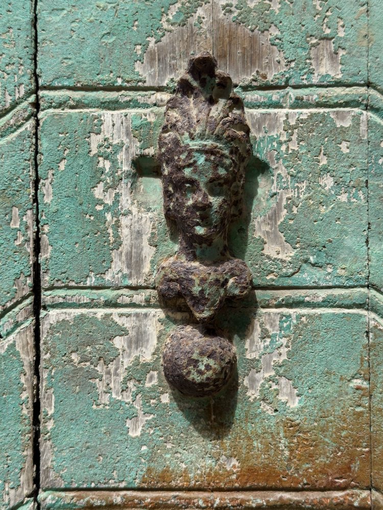 Metal Weathered Door Knocker shaped like a face with ornamental detail on a green door panel with peeling paint and rust, Scilla, Calabria, Italy.