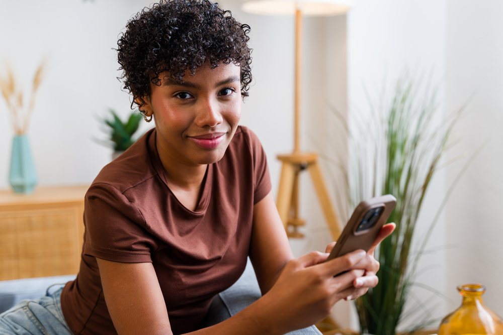 Young woman with curly hair in brown shirt sitting indoors holding a smartphone, Spain.